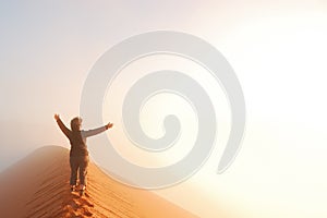 Person standing on top of dune in desert and looking at rising sun in mist with hands up, travel in Africa