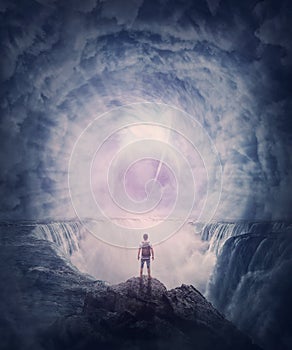 Person stands on the edge of a cliff above a waterfall looking at a huge whirlwind in the clouds that creates a portal to another