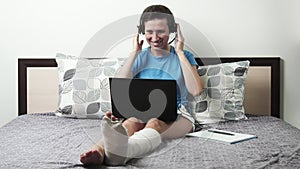 A person using a laptop computer sitting on top of a bed. Woman with a broken leg sitting on the bed communicates