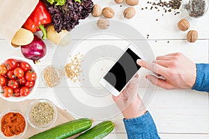Person using smartphone with blank screen while cooking and fresh ingredients on wooden table