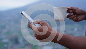 Person using smartphone. Hand of businessman checking phone while holding a cup of coffee on blue mountain background