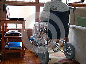 Person using a wheelchair, with a plaster cast on her leg, lloking through the window..Small desk with a laptop close to her. photo