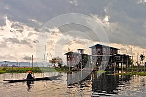 A person wearing a conical hat in a small boat rowing towards a stilted house during sunset at the Inle Lake in Myanmar.