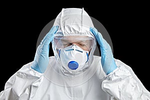 Person in white protective suit, facepiece respirator and glasses. Man holds his head, hands in disposable blue gloves