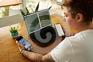 Person at wooden table multitasks with coding on laptop, analyzes data on two smartphones. Concentrated individual with