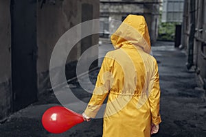 Person in yellow cloak with hood with red balloon going away to the dark old courtyard