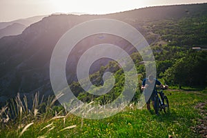 Perspective of a fit mountain biker pushing his bike uphill with amazing view on a forest, river and mountains in the background.