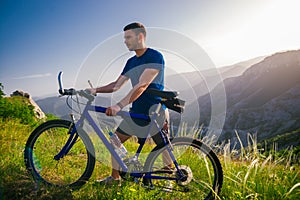 Perspective of a fit mountain biker pushing his bike uphill with amazing view on a forest, river and mountains in the background.