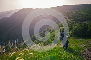 Perspective of a fit mountain biker pushing his bike uphill with amazing view on a forest, river and mountains in the background.