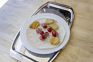 Perspective shot of chicken nuggets and small red fresh tomatoes on the wide white plate as a breakfast with thin bread on metal t