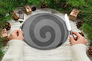 Perspective view girl holds fork and knife in hand and is ready to eat. Empty black slate plate on wooden christmas background. ho