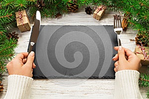 Perspective view girl holds fork and knife in hand and is ready to eat. Empty black slate rectangular plate on wooden christmas