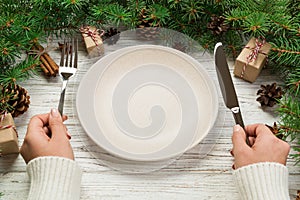 Perspective view girl holds fork and knife in hand and is ready to eat. Empty plate round ceramic on wooden christmas background.