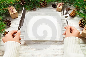 Perspective view girl holds fork and knife in hand and is ready to eat. Empty white square plate on wooden christmas background.