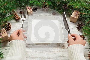 Perspective view girl holds fork and knife in hand and is ready to eat. Empty white square plate on wooden christmas background. h