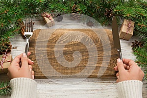 Perspective view girl holds fork and knife in hand and is ready to eat. Empty wood rectangular plate on wooden christmas backgroun