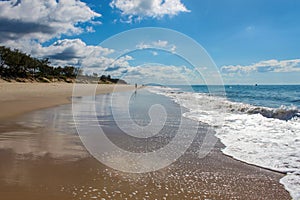 Perspective of wet beach reflecting blue cloudy sky with waves coming in and sihlouttes of people walking toward the horizon and