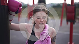 Perspiring beautiful woman in boxing gloves rubbing sweat off forehead looking away. Portrait of tired Caucasian