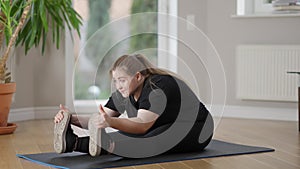 Perspiring overweight Caucasian sportswoman sitting on exercise mat bending forward. Portrait of motivated persistent