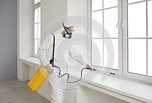 Pest control worker in protective suit and mask spraying chemical over house window