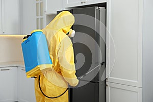 Pest control worker in protective suit spraying insecticide near refrigerator