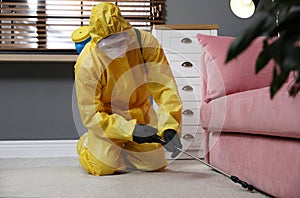 Pest control worker in protective suit spraying insecticide under sofa