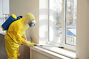 Pest control worker in protective suit spraying pesticide near window