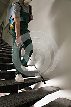 Pest control worker in uniform spraying pesticide on stairs indoors