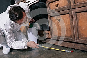 pest control worker in uniform spraying pesticides under cabinet