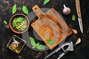 Pesto sauce, empty cutting board, basil leaves, garlic and olive oil on a dark rustic background. View from above