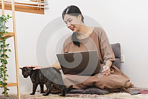 Pet lover concept, Young Asian woman using laptop and playing with the cat in living room at home