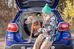 Pet owner woman in road trip holding magyar vizsla dog on leash sitting in car trunk on nature.