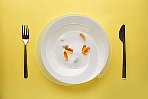 Pharmaceutical medicine pills or capsules on a plate with a fork and knife. Top view yellow background