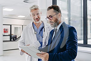 Pharmaceutical sales representative talking with doctor in medical building, presenting new medication on tablet