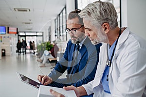 Pharmaceutical sales representative talking with doctor in medical building, presenting new medication on tablet