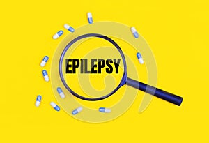 Pharmaceutical tablets and capsules near a magnifying glass with the text EPILEPSY on a yellow background. View from above