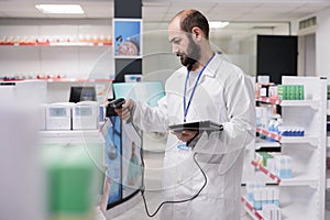 Pharmacist in white coat holding tablet computer typing products barcode while doing pills inventory
