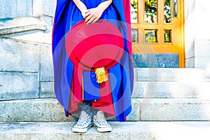 PhD doctoral graduate in regalia gown, holding tudor bonnet cap, sitting on university steps, with sneaker canvas shoes showing