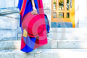 PhD doctoral graduate in regalia gown, holding Tudor bonnet cap, sitting on university steps, with sneaker canvas shoes showing photo