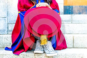 PhD doctoral graduate in regalia gown, holding tudor bonnet cap, sitting on university steps, with sneaker canvas shoes showing