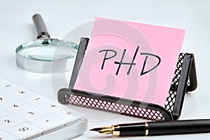 PHD word on the sticker on the stand on a white background next to a fountain pen, calculator and magnifying glass on a white
