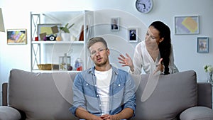 Phlegmatic man sitting on sofa listening nervous wife shouting, aggression