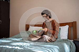 Teen girl reading messages scrolling social media in smartphone drinking tea in bed. Phone addiction