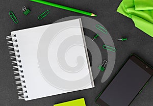 Phone with blank white spiral paper notebook, colored pencil, paper clips, paper clumps and green cloth on dark background