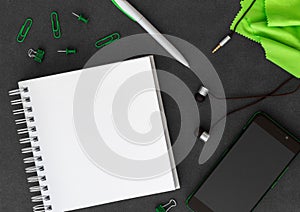 Phone with blank white spiral paper notebook, pen, paper clips, paper clumps, headphones and green cloth on dark background