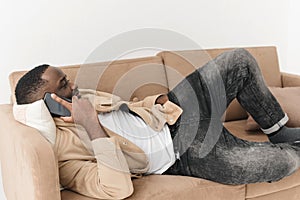 Phone call. Happy afro man talking on mobile phone while lying on sofa at home.