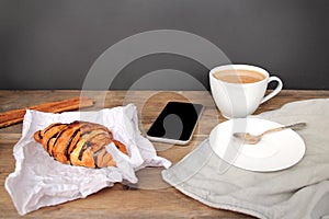 Phone and cappuccino coffee in a white cup and saucer and chocolate muffin in paper on an old wooden table, coffee maker, food