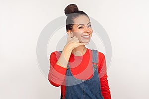 Phone conversation! Portrait of positive stylish pretty girl with hair bun in denim overalls showing call me gesture