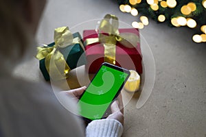 A phone with a green screen in the hands of a woman in a white sweater, a red and green gift box and bokeh lights on the