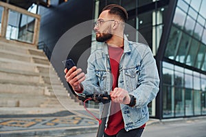 With phone in hands. Handsome young guy in casual clothes standing with electric schooter outdoors at sunny daytime